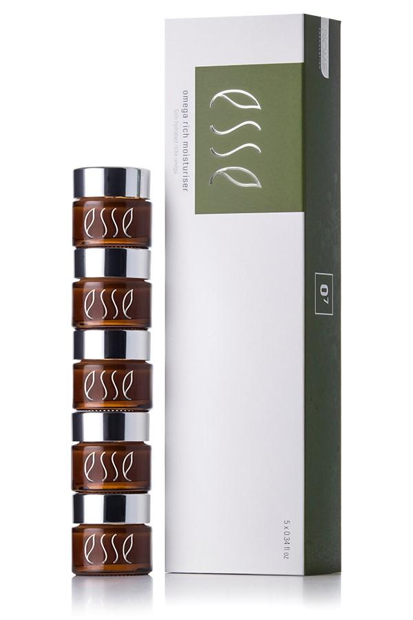A stack of five small brown jars with silver lids labeled "Esse Omega Rich Moisturizer" is placed next to a tall white and green box that also displays the same product name. The jars are aligned vertically, and the box stands upright on the right side of the image.