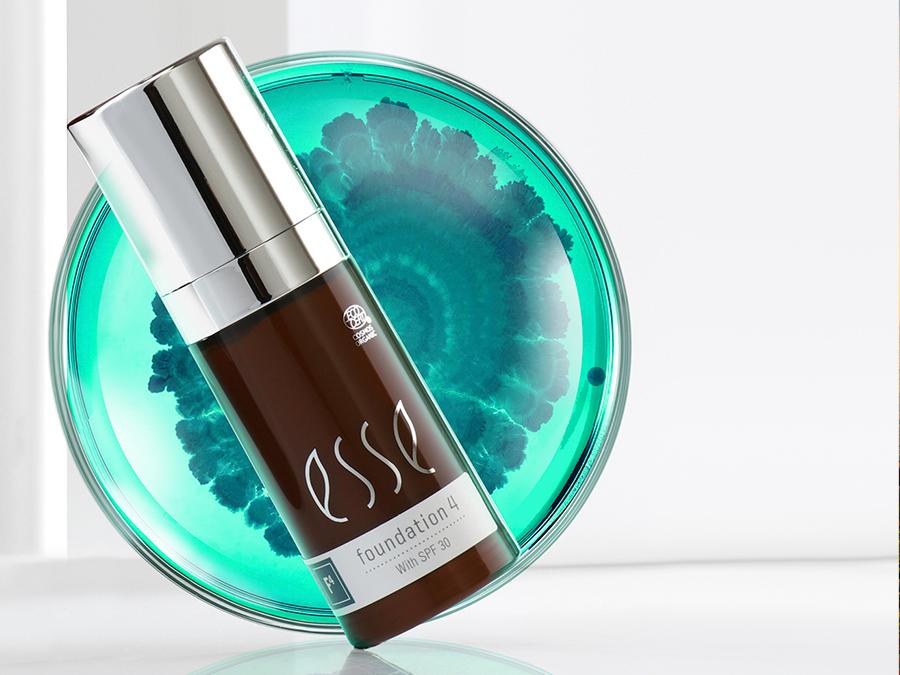 A bottle of Esse Concealer with SPF 30 stands in front of a round, artistic background resembling a turquoise and blue abstract floral pattern. The concealer bottle has a silver cap and a dark brown base with white text.