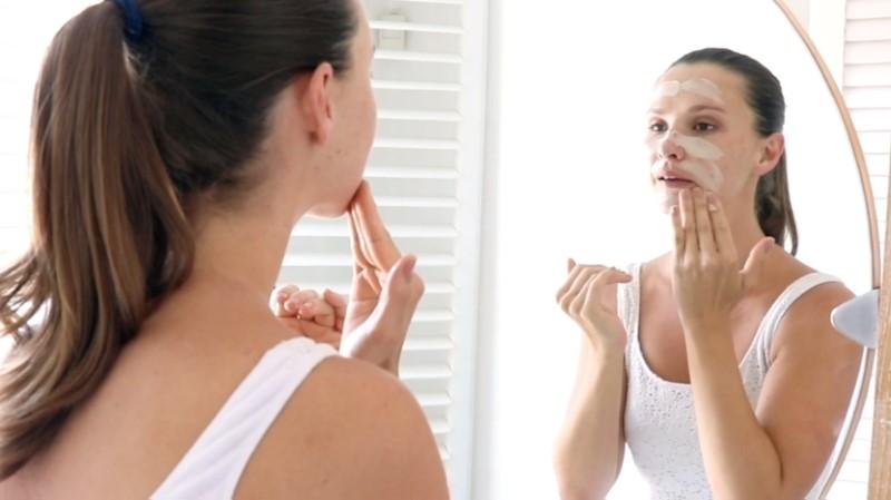 A woman with brown hair, tied back in a ponytail, stands in front of a mirror applying the Esse Clay Mask to her face. Dressed in a white tank top, she uses her fingers to evenly spread the mask. The bright background features white shutters.