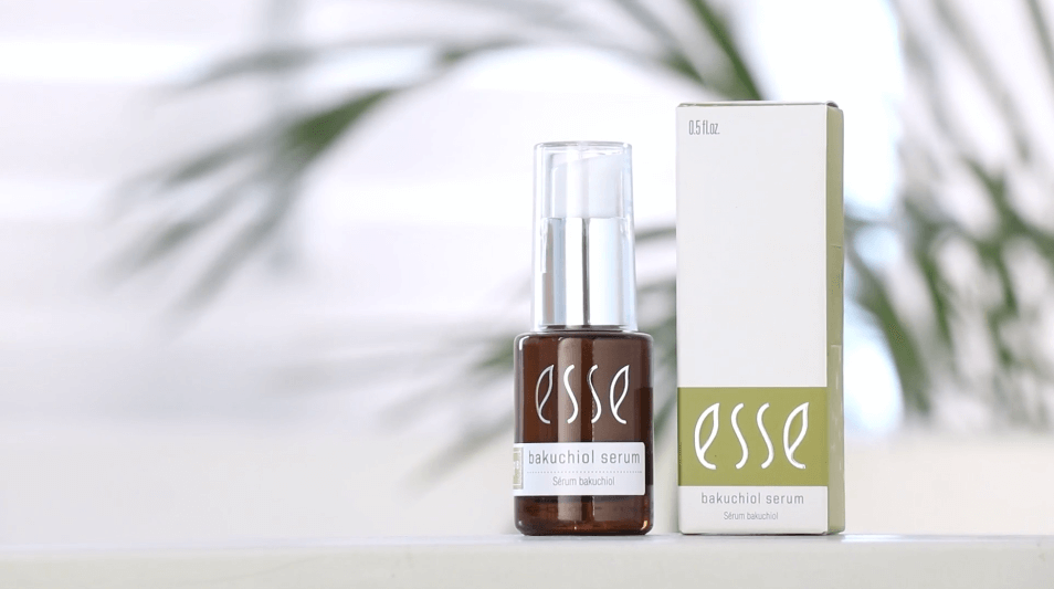 A brown bottle adorned with the label "Esse Bakuchiol Serum" and equipped with a silver pump dispenser stands beside its coordinating white and green packaging box. The softly blurred background, illuminated by natural light, features subtle hints of green foliage.