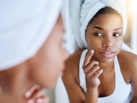 A woman wrapped in a towel turban examines her face closely in the mirror, gently touching her cheek with one finger. She appears thoughtful, focusing on her skin in a well-lit bathroom setting.