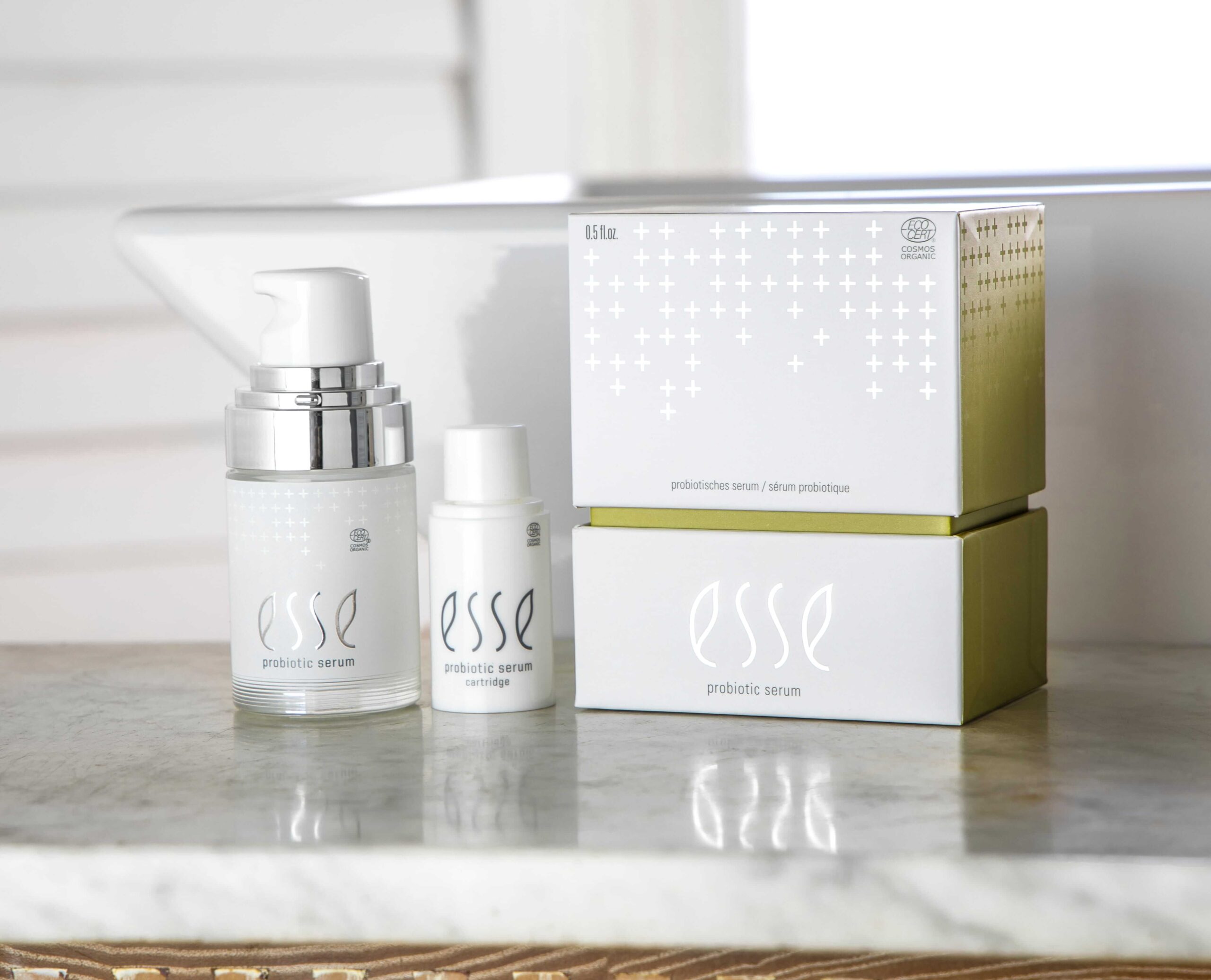 Two bottles of Esse Probiotic Serum—one with a pump and a smaller one—are shown on a marble surface alongside their white and green boxes, with a window and white paneling visible in the background.
